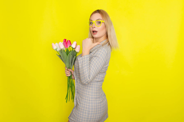 Blonde woman in plaid dress holding tulips bouquet over yellow background - Photo, Image