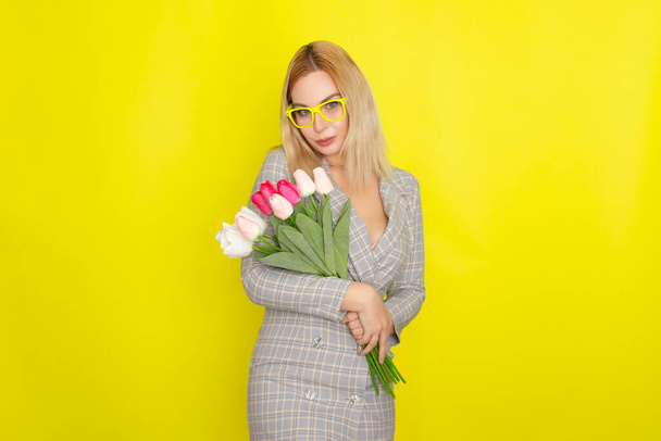 Blonde woman in plaid dress holding tulips bouquet over yellow background - Фото, зображення