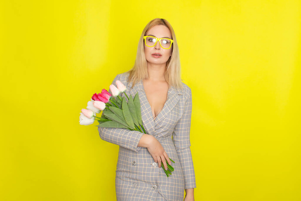 Blonde woman in plaid dress holding tulips bouquet over yellow background - Fotó, kép