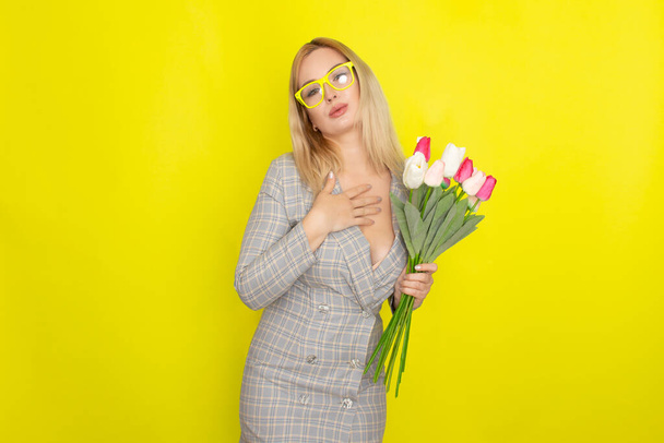 Blonde woman in plaid dress holding tulips bouquet over yellow background - Photo, image