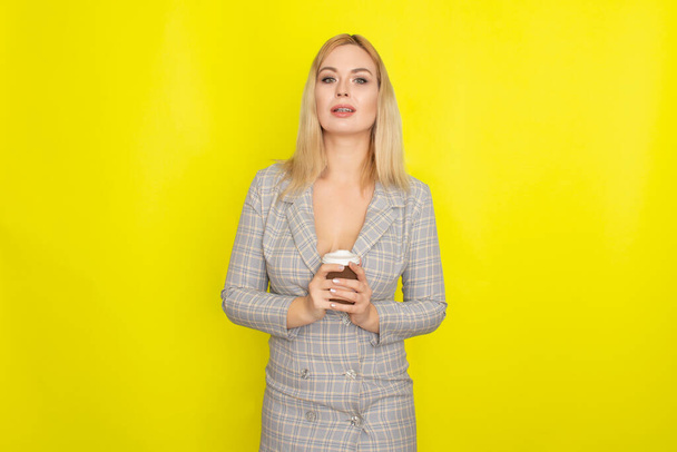 Business blonde woman wearing plaid jacket style dress and drinking coffee over yellow background - Fotografie, Obrázek
