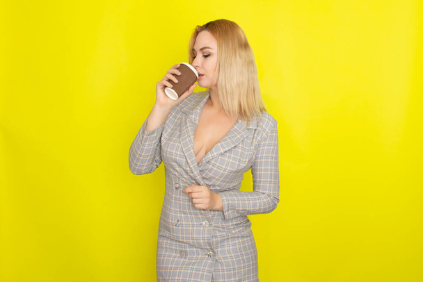 Business blonde woman wearing plaid jacket style dress and drinking coffee over yellow background - Фото, зображення
