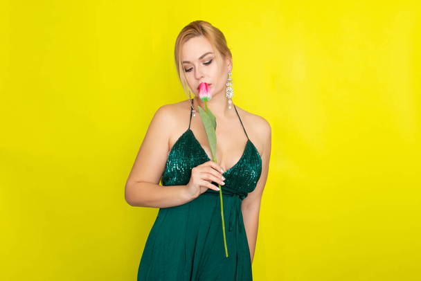 Blonde woman in green evening dress holding one tulip in her hands over yellow background - Foto, Imagen