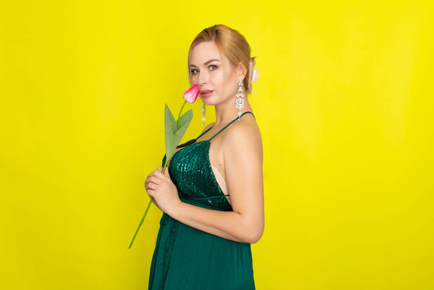 Blonde woman in green evening dress holding one tulip in her hands over yellow background - Foto, imagen