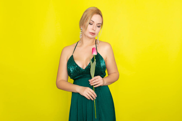 Blonde woman in green evening dress holding one tulip in her hands over yellow background - Fotó, kép