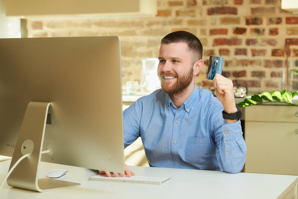Ein glücklicher Mann mit Bart sitzt vor dem Computer und wählt zu Hause in einem Online-Shop Produkte aus. Ein Mann macht eine Online-Zahlung im Internet auf einem Desktop-Computer in seiner Wohnung. - Foto, Bild