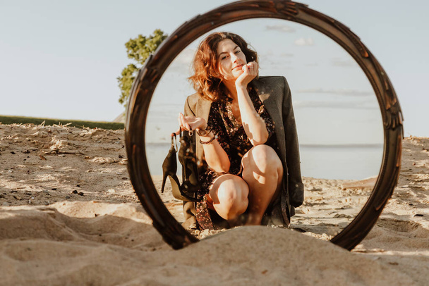 Retrato de estilo de vida de moda de la joven mujer de moda vestida con vestido negro y chaqueta riendo, posando en la playa en el reflejo del espejo; en el fondo arenoso y el mar
 - Foto, Imagen