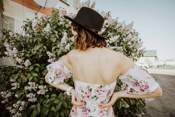 Szépség nő egy romantikus fény ruha csupasz vállak és a kalap egy tavaszi kertben virágzó orgonák - Fotó, kép