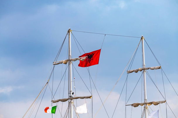 Maszt zabytkowego masztu drewniany żaglowiec na wycieczki morskie w porcie Saranda, Albania z czerwonym stanem albański flaga z czarnym dwugłowym orłem, i flaga włoska. - Zdjęcie, obraz