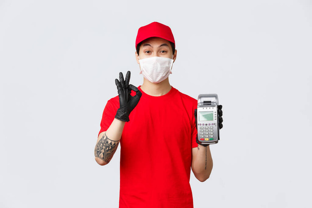 Contactless delivery, safe purchase and shopping during coronavirus concept. Friendly asian courier in red uniform cap and t-shirt, wear medical mask and gloves, show POS terminal and okay sign - Photo, Image