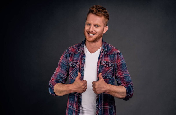 Nice job man, like it.Pleased good-looking happy male friend with ginger hair and brislte, showing thumbs up and smiling broadly, giving positive feedback, sharing his positive opinion over grey wall. - Foto, Imagem