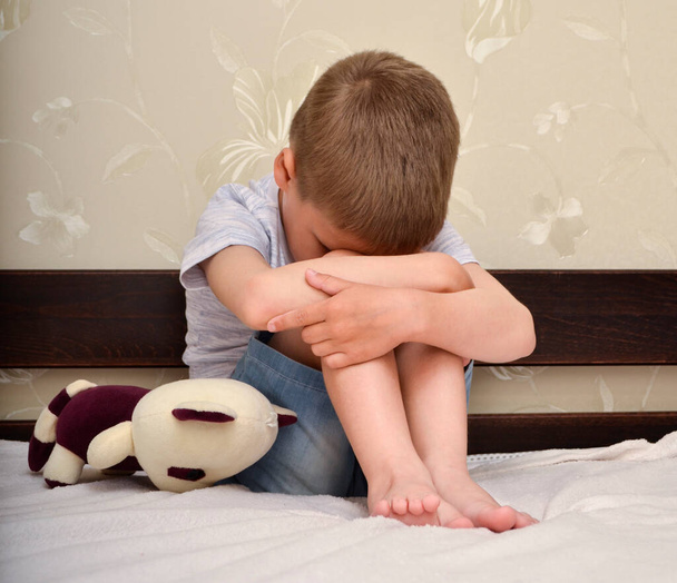 子供は両手で顔と足を組んでベッドの上に座っている。家庭内暴力。児童虐待. - 写真・画像