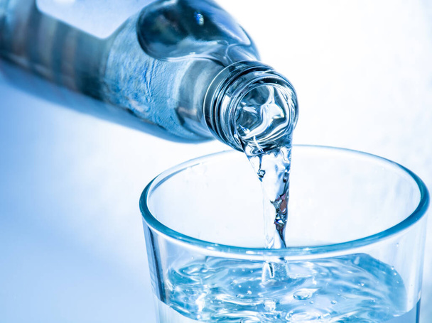 Gieten van koud helder drinkwater uit fles in glas, close-up, blauw getint op witte achtergrond - Foto, afbeelding