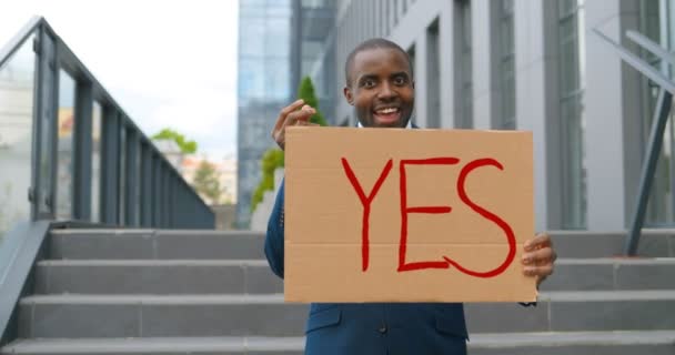 Portré felvétel afro-amerikai férfi kezében poszter Igen, és csinál pénzt gesztus demonstráció vagy tiltakozás. Egy tiltakozás a szabadban. Aktivista koncepció. Vidám ember mutatja asztal szavakkal igen. - Felvétel, videó