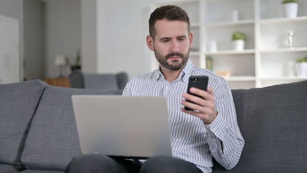 Uomo concentrato che lavora su smartphone e laptop a casa
  - Foto, immagini