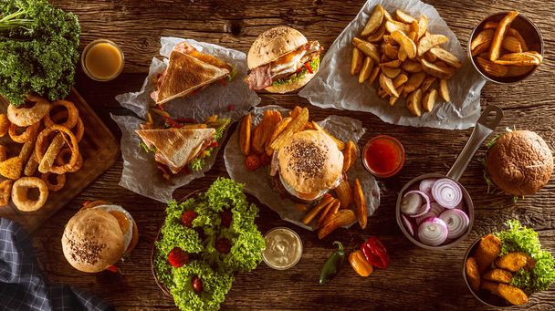 Top view of a table full of fast food, including hamburgers, potato chips, club sandwich, chicken nuggets and onion rings on a rustic table - Foto, Imagen
