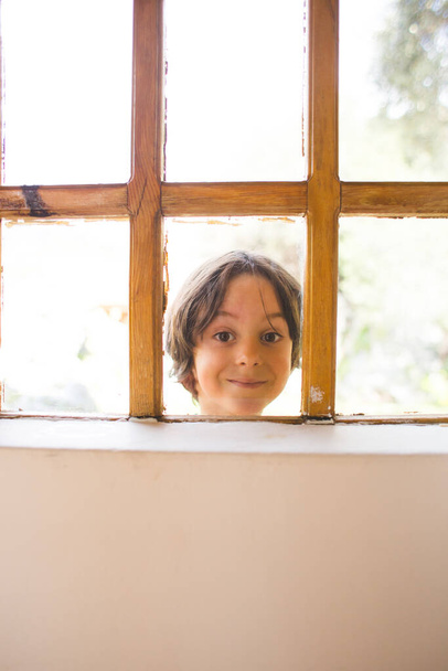 Egy kíváncsi fiú néz ki az ablakon, egy gyerek kukucskál be a házba az ablakon, játékos gyerek, portré egy barna szemű gyerekről, Megfigyelés az ablakon keresztül, egy gyerek érzelmei. - Fotó, kép