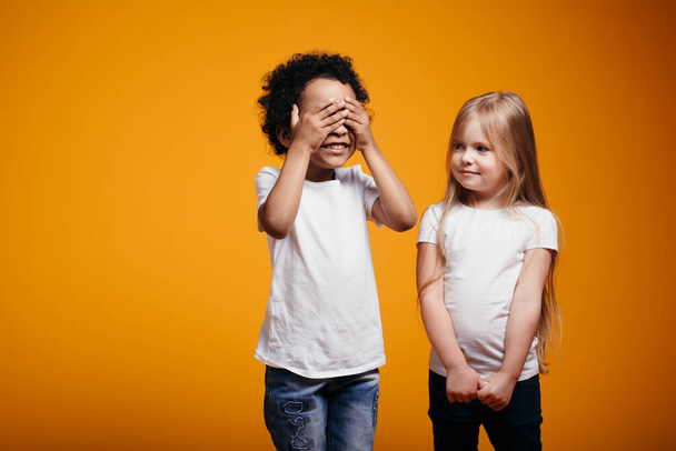 The dark-skinned boy closed his eyes and considers to start playing hide and seek with a fair-haired girl on an orange background. - Fotografie, Obrázek