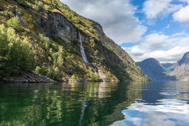 Cascada cayendo, norte fiordo Aurlandsfjord espejo de agua reflexión. Brillante vista de las montañas salvajes de la naturaleza de Noruega. Día de viaje soleado
. - Foto, Imagen