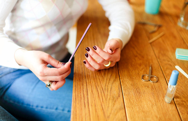 High angle side view of crop anonymous female in casual clothes sitting at wooden table with various tools for manicure and polishing nails with nail file - Foto, Imagem