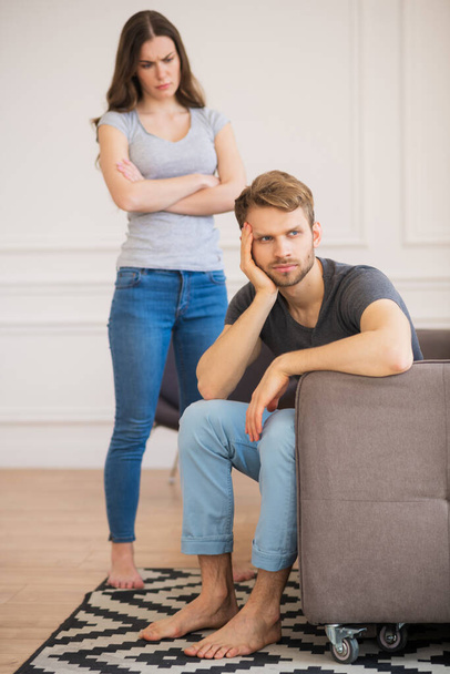 Young woman standing with arms clasped while her husband sitting with sad look - 写真・画像