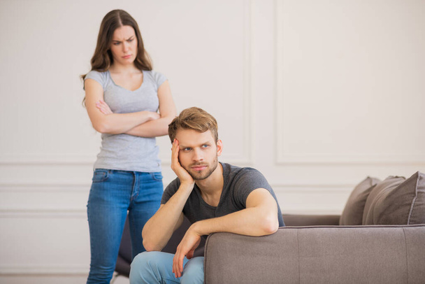 Young woman standing with arms clasped while her husband sitting with depressed look - Foto, afbeelding