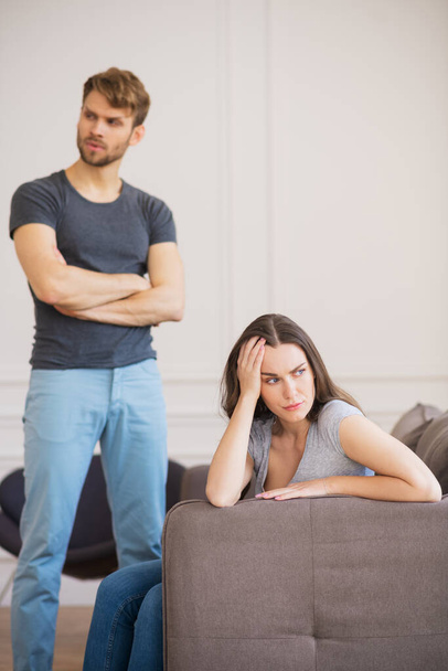 Young bearded man standing with arms clasped while his wife sitting with depressed look - Foto, Imagen