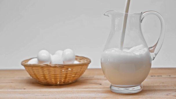 Zeitlupe der Milch, die in Kannen in der Nähe von Eiern auf einer auf grau isolierten Holzoberfläche gegossen wird  - Filmmaterial, Video