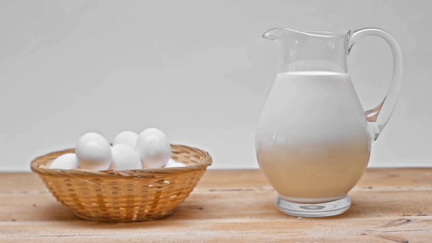 Vejce v proutěném koši a džbánek mléka na dřevěném povrchu izolované na šedé - Záběry, video