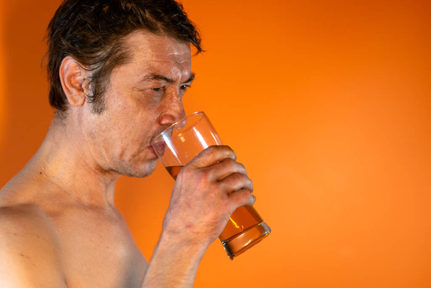 An adult male with a naked torso drinks beer from a glass glass. Concept: Alcoholism and the alcoholic man - Photo, Image