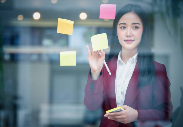 Portrét mladé asijské podnikatelky, která píše lepicí papír na sklo v moderní kanceláři. Koncept obchodního plánu. - Fotografie, Obrázek