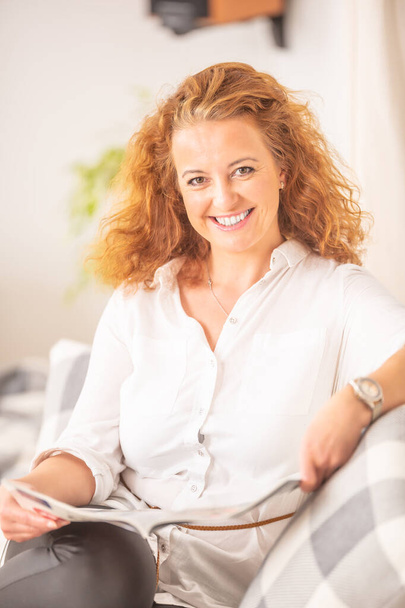 Femme aux cheveux bouclés lit un magazine, assise sur un canapé dans un salon, souriant à la caméra
. - Photo, image