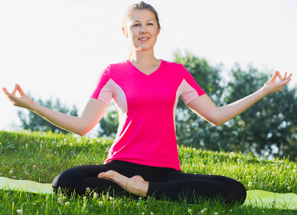 Female in pink T-shirt is sitting and doing meditation in the park. - Photo, Image