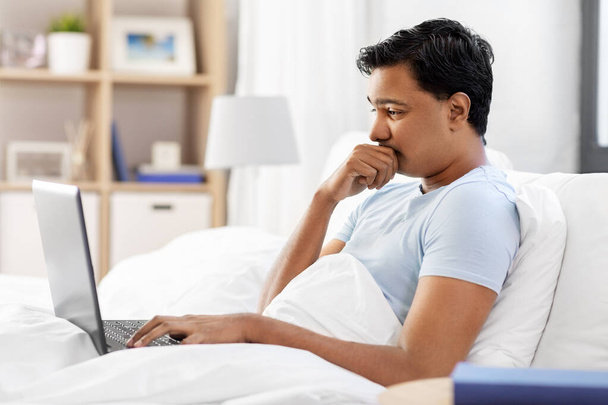 worried indian man with laptop in bed at home - Foto, afbeelding