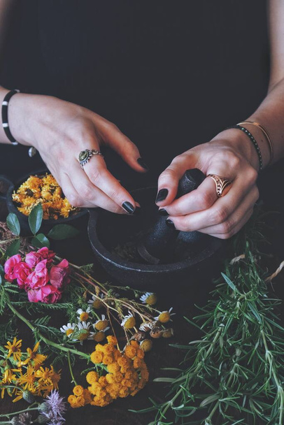 メスの魔女は夏のハーブや花を害虫やモルタルで研削します。黒のテーブルの上にカラフルな新鮮な花やローズマリー。手にはヴィンテージジュエリーを持った白人女性が杵を握っている。垂直画像 - 写真・画像