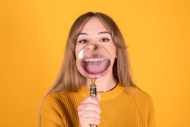 woman with a magnifying glass putting in front of her mouth, smile, isolated on yellow background - Photo, Image