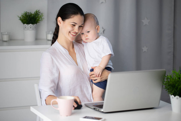 Young mother working in a decree at the computer with a baby son in her arms - Foto, Bild