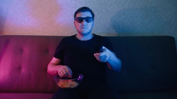 A man sits on a sofa in 3D glasses eating chips and switching channels on a TV. - Video
