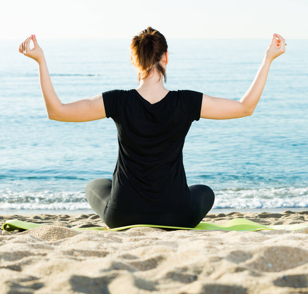 Portrait of sportswoman which is sitting her back and practicing asana in black T-shirt on the beach. - Foto, Imagem