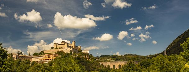 Blick auf Spoleto, grüne Berge, blauer Himmel mit weißen Wolken. Die Festung Rocca Albornoziana wird im Sommer von der Sonne erleuchtet. Die Brücke der Türme, römisches Aquädukt. Bäume im Vordergrund - Foto, Bild