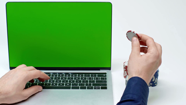 przycięty widok człowieka w pobliżu laptopa z zielonym ekranem trzymając żeton pokera na białym - Materiał filmowy, wideo