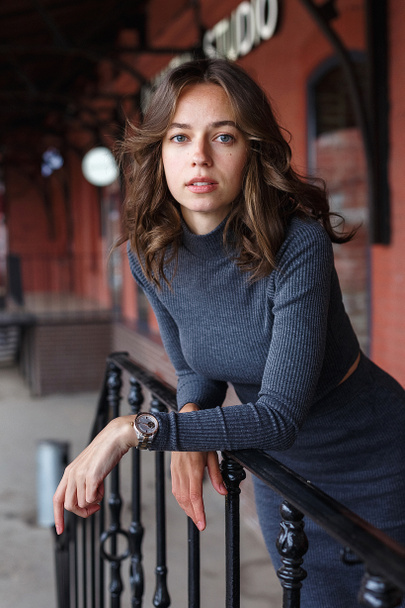 young thoughtful pretty girl in gray turtleneck, skirt with watches on arm poses in front of red building. Street style portrait photosession of attractive female, urban photoshoot of elegant model - Photo, Image