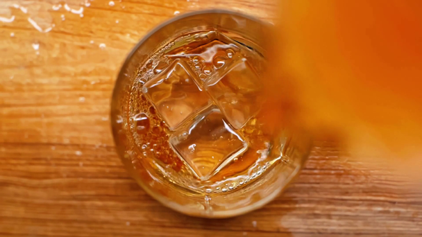 top view of ice cubes falling into glass and pouring whiskey on wooden table - Imágenes, Vídeo