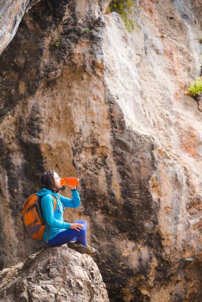 A woman with a backpack drinks water from a reusable bottle. A girl sits on a large stone and quenches her thirst. Traveler on the background of rocks. - Photo, Image