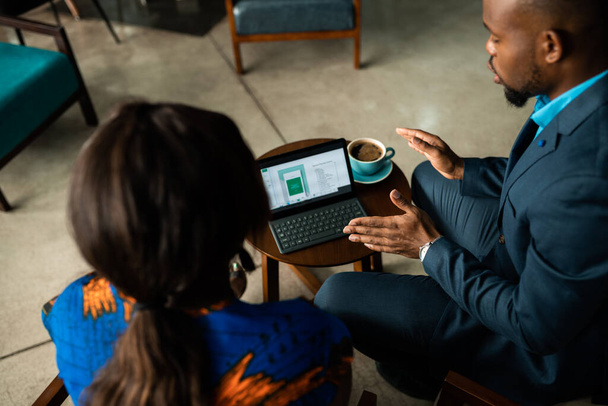 Twee jonge Afrikaanse zakenmensen praten over werk op een digitale tablet tijdens een informele bijeenkomst samen koffie drinken in een kantoor - Foto, afbeelding