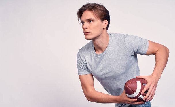 A young athletic Caucasian guy, with blond hair, in a gray basic t-shirt, holds an American football ball in his hands, prepares to make a throw. Copy space. - Foto, Imagem