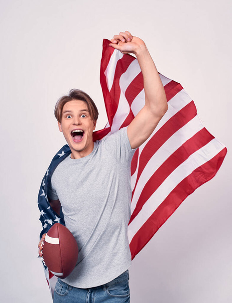Vertical shot, a young slender guy, in casual clothes, holds the American flag and ball for American football, runs and is smiling, screaming with joy. Emigrant concept, American symbolism, July 4th. - Foto, Imagen