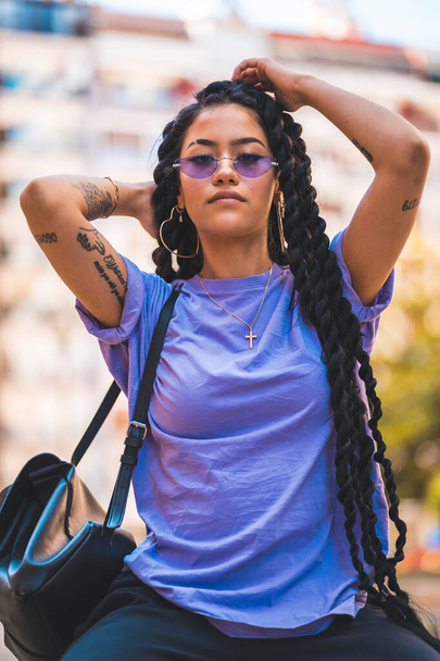Lifestyle, young dark-skinned woman with long braids and purple glasses sitting on a bench in the park with her backpack slung over her shoulder - Foto, immagini