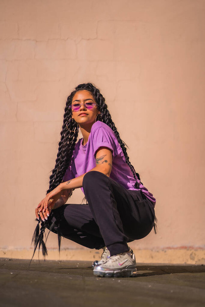 A dark-skinned young woman with long braids and purple glasses. In an urban shoot, on the ground with a smooth wall in the background - 写真・画像