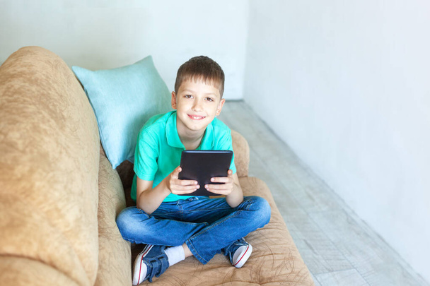 Child boy using digital tablet on sofa in living room. Kid studying from home and playing with gadget - Fotó, kép